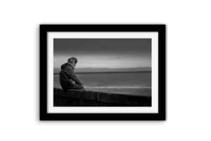 Femme assise seule sur un muret de pierres, penchée sur son téléphone, face à l'océan sous un ciel couvert — photo noir et blanc prise sur la plage de Bonne Source à Pornichet.