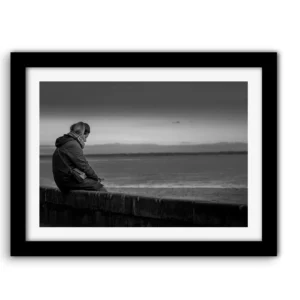 Femme assise seule sur un muret de pierres, penchée sur son téléphone, face à l'océan sous un ciel couvert — photo noir et blanc prise sur la plage de Bonne Source à Pornichet.