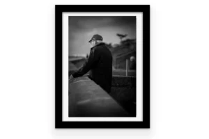 Homme âgé de dos, casquette et veste noire, appuyé sur un muret de granit face à l'océan sur le sentier côtier de Pornichet, sous un ciel nuageux — photo noir et blanc.