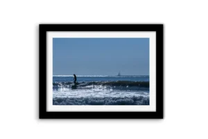 Silhouette en stand up paddle luttant contre les vagues sur la plage des Libraires à Pornichet, voilier et éoliennes en mer visibles à l'horizon sous un ciel bleu.