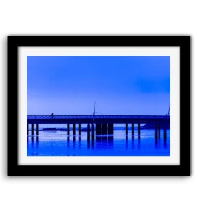 Silhouette solitaire traversant le pont du port de Pornichet, traité en nuances de bleus intenses, avec reflets des piliers dans l'eau calme et horizon lumineux.